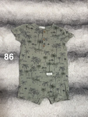 Grön byxdress med palmmönster Newbie - Kortärmad grön byxdress från Newbie med tryck av palmer och krokodiler. Knäppning med träknappar framtill och tryckknappar i grenen. perfekt för varma dagar.🌸 Fint använt skick. Storlek 86