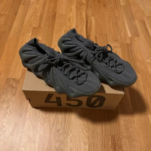 Adidas Yeezy 450 mörkgrå sneakers - Säljer ett par Adidas Yeezy 450 sneakers i mörkgrå färg. Skorna har en unik, vågig sula och stickad ovandel med snörning. Modellen har en futuristisk design och är tillverkad i syntet och textil för en skön passform. Perfekt för dig som gillar statement-sneakers. Sitter mindre än 45 1/3, sitter mer som 44. Helt nya, endast använda en gång. Lite svåra att få tag på eftersom adidas x yeezy samarbetet nu är avslutat och inget nytt säljs. Nypris låg på 1500kr, köpta från adidas hemsida.