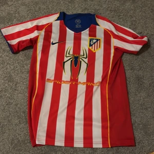 Atletico Madrid Spider-Man 2 fotbollströja - Säljer en Atletico Madrid fotbollströja från Nike med röda och vita ränder, blå detaljer och gula inslag. Framsidan har Spider-Man 2-tryck och klubbmärke, baksidan har F. Torres och nummer 9 i gult. Kortärmad och tillverkad i polyester.