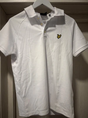 Vit pikétröja från Lyle & Scott - Vit pikétröja från Lyle & Scott med klassisk krage, knappar framtill och det ikoniska gula fågel-loggan på bröstet. Tröjan har en snygg ribbad detalj längs ena sidan framtill och är tillverkad i mjuk bomull. Perfekt för en clean och sportig look.