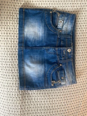 Blå jeanskjol med hjärt-detaljer - Säljer en blå jeanskjol i kort modell med snygga slitningar och coola detaljer som dragkedjor med hjärtformade hängen. Kjolen har flera fickor och dekorativa nitar, perfekt för dig som gillar en trendig och ungdomlig stil. Den är använd 1 gång! En knapp borta