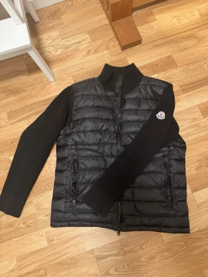 Svart Moncler cardigan med stickade ärmar - Säljer en svart cardigan från Moncler med stickade ärmar och rygg. Jackan har två dragkedjefickor fram, hög krage och klassisk Moncler-logga på ärmen. Framsidan är quiltad och fodrad, medan ärmar och baksida är ribbstickade för extra komfort.