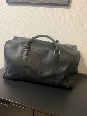 Svart weekendbag från Valentino - Säljer en stilren svart weekendbag från Valentino i skinnimitation. Väskan har dubbla handtag, silverfärgade metalldetaljer och dragkedja upptill. Perfekt storlek för övernattning eller resa och har en clean look med diskret logga på framsidan.. ALDRIG använd! Nypris 2,5k