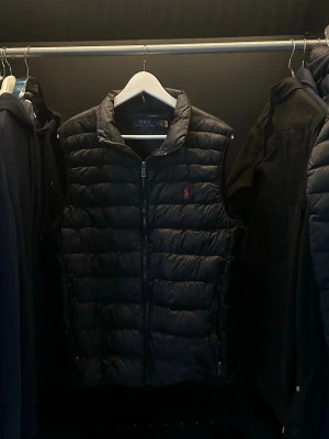 Svart dunväst från Polo Ralph Lauren - Säljer en svart dunväst från Polo Ralph Lauren med quiltad design och dragkedja framtill. Västen har den klassiska röda Polo-loggan broderad på bröstet och två fickor med dragkedja. Perfekt att slänga över en hoodie eller tröja för en clean look.