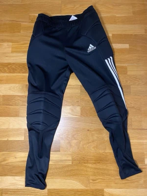 Svarta Adidas fotbollsbyxor M - Säljer ett par svarta Adidas fotbollsbyxor i storlek M. Byxorna har vadderade partier vid knän och höfter, samt klassiska vita ränder längs benen och Adidas-logga på låret. Tillverkade i polyester för maximal rörelsefrihet och komfort.