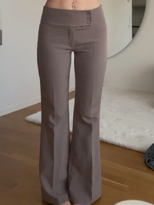 Taupe bootcut kostymbyxor med knappar - Säljer ett par bruna kostymbyxor med medelhög midja och bootcut passform. Byxorna har bred linning med tre dekorativa knappar i sidan och pressveck framtill. Materialet är slätt och ger en clean look. Perfekta för dig som gillar en stilren vibe.
