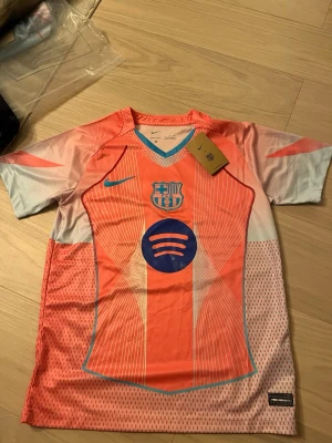 Barça x Nike fotbollströja rosa/blå - Säljer en Barcelona x Nike fotbollströja i orange och ljusblå med coola grafiska linjer och Spotify-logga på bröstet. Tröjan har FCB-emblem, Dri-Fit material och V-ringad krage med blå kant. Perfekt för dig som älskar fotboll och vill sticka ut på planen.