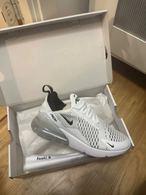 Nike Air Max 270 vita sneakers helt nya  - Nike Air Max 270 sneakers i vitt med svarta detaljer och stor synlig luftbubbla i hälen. Ovandelen är i mesh med ventilerande hål och snörning. Snygg och sportig design som sticker ut med sin futuristiska vibe och bekväma passform. Helt nya 