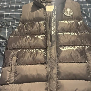 Svart dunväst från Moncler - Säljer en svart dunväst från Moncler med hög krage och tydlig logga på bröstet. Västen har en quiltad design, dragkedja framtill och stora fickor. Insidan har en unik serietidningsdetalj. Perfekt för lager-på-lager och streetwear.