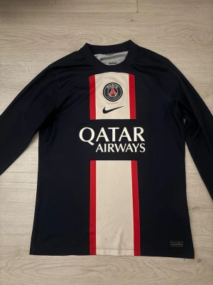 PSG långärmad matchtröja Nike - Säljer en snygg långärmad Paris Saint-Germain matchtröja från Nike. Tröjan är marinblå med vit och röd randig design på framsidan, PSG-logga och Qatar Airways tryck. Perfekt för dig som älskar fotboll och vill ha en stilren supportertröja.