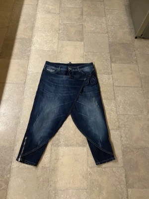 Unika jeans med omlott från SixtyDays - Säljer ett par mörkblå jeans från SixtyDays i omlottmodell med snörning i midjan och dragkedjor vid bensluten. Jeansen har croppad längd och coola slitningar framtill. Materialet är bomull med lite elastan för extra komfort.