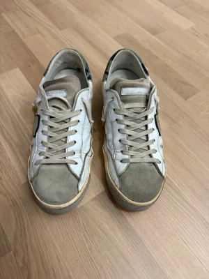 Philippe Model  - Säljer ett par vita Philippe Model sneakers med beige detaljer och grå mocka på tån. Skorna har snörning, en broderad sköld på sidan och inslag av blått på hälen. Tillverkade i skinn och mocka för en schysst premiumkänsla.