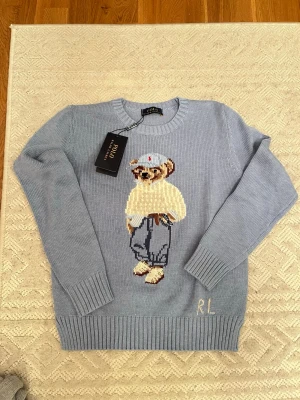 Blå stickad tröja Polo Ralph Lauren - Säljer en ljusblå stickad tröja från Polo Ralph Lauren med ikonisk björn på bröstet. Tröjan har rund halsringning, ribbade muddar och RL-broderi nedtill. Perfekt för dig som vill ha något klassiskt men ändå lekfullt.