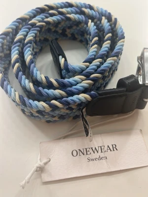 Blå- och beige flätad bälte ONEWear - Säljer ett snyggt flätat bälte från ONEWear i olika blå nyanser och beige. Bältet har detaljer i svart skinn och metallspänne. Perfekt accessoar för att lyfta din outfit med en cool twist.