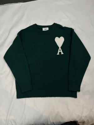 Grön stickad tröja från Ami Paris - Mörkgrön stickad tröja från Ami Paris med rund hals och ribbade muddar. På bröstet finns ett vitt hjärta och bokstaven A som snygg detalj. Perfekt för dig som gillar stilrena plagg med en twist. Materialet är mjukt och skönt.