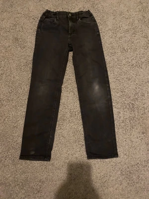 Svarta jeansbyxor från Lindex, strl 152 - Svarta jeansbyxor från Lindex i storlek 152. Klassisk femficksmodell med rak passform och resår i midjan för extra komfort. Snyggt slitna detaljer och knappstängning framtill. Perfekt för dig som gillar enkel och stilren look.