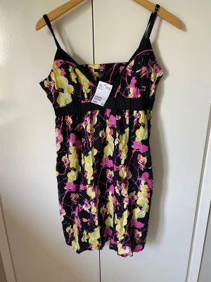 Blommig klänning  - Superfin klänning från H&M med svart botten och färgglada blommor i rosa och gult. Klänningen har smala axelband, v-ringning och markerad midja. Perfekt för varma dagar när du vill sticka ut lite extra.