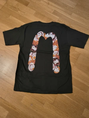 Svart Evisu t-shirt med grafiskt tryck - Svart t-shirt från Evisu med färgstarkt grafiskt tryck på bröstet och stor M-logga med japanskt inspirerade figurer på ryggen. Klassisk rund hals och korta ärmar. Materialet är mjuk bomull som känns skön mot huden. Perfekt för dig som gillar streetwear med unik design.