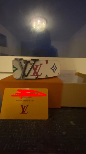 L bälte  - Säljer ett snyggt vittL bälte med ikoniskt L-spänne i metall. Bältet har färgglada loggor och symboler i rött, blått, grönt och rosa längs hela bandet. Perfekt accessoar för att lyfta din outfit med lyxig vibe. Kommer med kvitto och box kom med bud