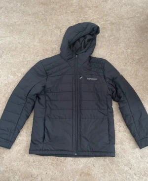 Svart pufferjacka Peak Performance - Svart pufferjacka från Peak Performance med huva och diskret logga på bröstet. Jackan har quiltad design, dragkedja framtill och två sidofickor. Perfekt för kalla dagar och har en clean, sportig look som funkar till det mesta.