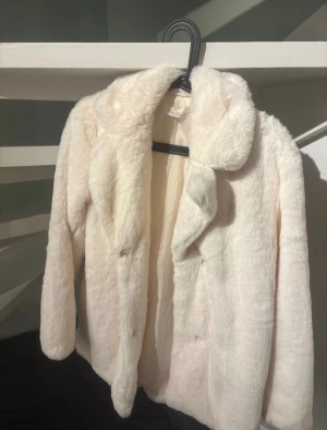 Vit pälsjacka från H&M - Säljer en fluffig och mjuk vit pälsjacka från H&M med bred krage och långa ärmar. Jackan har en clean look och stängs med dolda knappar framtill. Perfekt för dig som vill ha en cozy och stylish vibe under kyliga dagar.