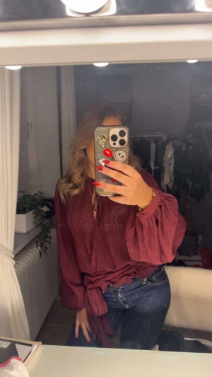 Vinröd plisserad blus med knyt - Säljer en vinröd plisserad blus med långa, vida ärmar och knyt i midjan. Blusen har en lätt transparent känsla och snygga detaljer vid ärmsluten. Perfekt för dig som gillar trendiga och unika plagg med lite extra volym. Kommer inte till användning