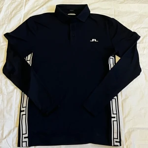 Mörkblå långärmad skjorta J.Lindeberg - Snygg mörkblå långärmad skjorta från J.Lindeberg med vit logga på bröstet och ryggen. Skjortan har krage, knappar framtill och coola vita grafiska detaljer längs sidorna. Perfekt för dig som gillar stilren design med sportig touch.