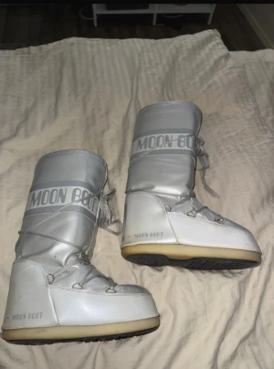 Silvriga Moon Boots med snörning - Moon Boots i metallic silver med ljusgrå detaljer och snörning runt hela skaftet. De har en rund tå, platt sula och är fodrade för extra värme. Perfekta för vintern och snöiga dagar, med ikonisk logga på skaftet. Många defekter men inget som syns om man inte kollar riktigt noga, syns även inte ute i snön! Därav mitt pris även äldre model.