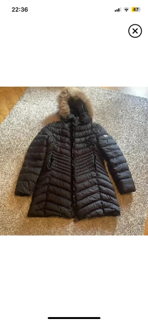 Svart Moncler dunjacka med päls - Svart lång dunjacka från Moncler med snygg päls på huvan och cool quiltad design. Jackan har tvåvägsdragkedja med Moncler-logga, långa ärmar och figurnära passform. Perfekt för kalla dagar med stil.
