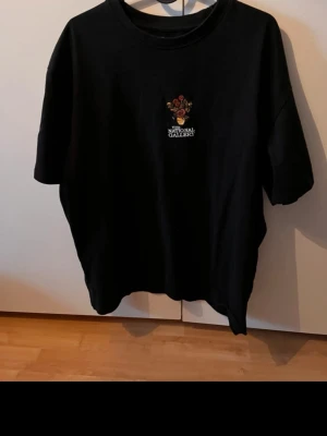 Svart t-shirt The National Gallery - Svart t-shirt med broderad blomdetalj och texten 'The National Gallery' på bröstet. Klassisk rund hals och korta ärmar, tillverkad i mjuk bomull. Passar dig som gillar konst och stilrena plagg med en twist.