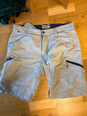 Beige McKinley shorts med dragkedja - Säljer ett par beige shorts från McKinley i storlek M. De har flera praktiska fickor, bland annat med dragkedja på sidorna, och bälteshällor. Materialet är lätt och passar perfekt för utomhusaktiviteter. Snyggt sportig look med diskreta detaljer.