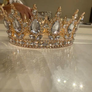Glittrig guld krona med kristaller - Säljer en riktigt maffig krona i guld med stora och små klara kristaller. Kronan har glittriga detaljer och spetsiga former som ger en kunglig vibe. Perfekt för bal, fest eller nyår. Kristallerna är infattade i ett guldfärgat metallband med snyggt mönster.