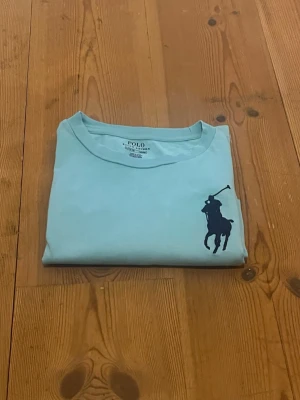 Ljusblå Polo Ralph Lauren t-shirt - Fräsch ljusblå t-shirt från Polo Ralph Lauren med klassisk stor mörkblå polospelare på bröstet och tryck på ena ärmen. Rund hals och korta ärmar, tillverkad i mjuk bomull. Perfekt för dig som gillar stilrena och sportiga plagg. 