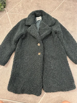 Svart teddyjacka från Gold & Silver - Mysig grön  teddyjacka från Gold & Silver med två knappar framtill och bred krage. Jackan har en fluffig, grov yta och är fodrad på insidan för extra komfort. Perfekt för dig som gillar oversized och chill stil.