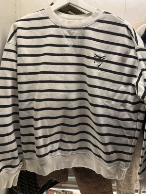 Randig sweatshirt med broderad detalj - Säljer en vit och svart randig sweatshirt med broderad rosett och text på bröstet. Tröjan har rund halsringning, ribbade muddar och en relaxed passform. Perfekt för dig som gillar klassisk stil med en söt twist.