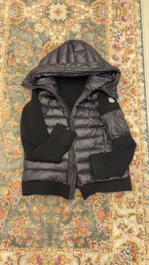 Svart Moncler cardigan med huva - Svart Moncler cardigan från Moncler med stickade ärmar och rygg, quiltad framsida och huva. Jackan har dragkedja, fickor med dragkedja och Moncler-logga på ärmen. Materialmix av polyester och stickad ull ger en cool kontrast. Perfekt för dig som gillar streetstyle.