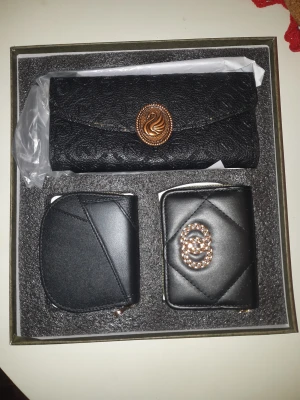 Set med svart plånbok och fodral - Snyggt set med svart plånbok, korthållare och myntfodral i skinnimitation. Plånboken har ett elegant mönster och guldigt emblem med svan, medan korthållaren har quiltad design och glittrigt dubbelring-emblem. Perfekt för att hålla koll på kort och pengar med stil.
