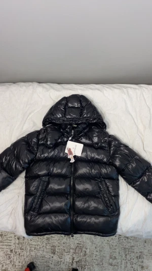 Svart Moncler pufferjacka med huva - Säljer en svart pufferjacka från Moncler med glansig finish och huva. Jackan har dragkedja framtill, två sidofickor med dragkedja och en ficka med Moncler-logga på ärmen. Perfekt för kalla dagar och har en riktigt snygg streetstil. Helt oanvänt och ren Pris kan sänkas