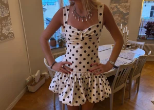 Prickig vit kortklänning med volang - Säljer en superfin vit kortklänning med svarta prickar och volang nedtill. Klänningen har breda axelband, söt rosettdetalj vid bysten och en figurnära passform upptill. 