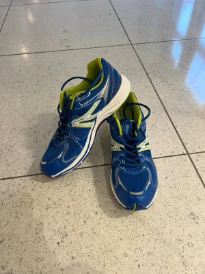 Blå sportiga sneakers med snörning - Snygga blå sneakers med vita och limegröna detaljer. Skorna har rund tå, platt sula och snörning. Ovandelen är i syntetmaterial med meshpaneler för extra ventilation. Insidan är limegrön och sulan är vit med blå och gröna inslag. Perfekta för träning eller en aktiv vardag.