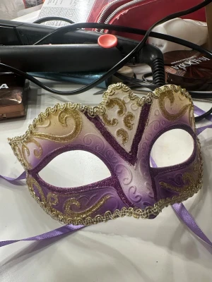 Lila och guld maskeradmask med glitter - Säljer en snygg maskeradmask i lila och vitt med glittriga detaljer i guld. Masken har vackra snirkliga mönster och en dekorativ kant i guld. Knyts med lila band bakom huvudet och passar perfekt till temafest eller bal.