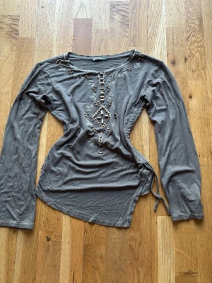 Brun/grön långärmad topp från I Heart Hemp - Supersnygg brun långärmad topp med broderade detaljer och pärlor framtill. Toppen har knytning i sidan och en unik asymmetrisk nederkant. Perfekt för dig som gillar boho-vibbar och coola detaljer.