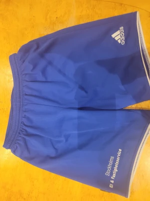 Blå Adidas shorts med snörning - Säljer ett par blå sportiga Adidas shorts med vit snörning i midjan och vit kant längs benen. De har tryck med 'Stockholms El & Fastighetsservice' på ena benet och är gjorda i lätt polyester med meshfoder. Perfekta för träning eller fotboll.