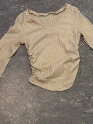 Beige ribbad topp med v-ringning - Säljer en beige ribbad topp med v-ringning och lång ärm. Toppen har snygg rynkning på ena sidan som ger en cool asymmetrisk look. Perfekt för dig som gillar neutrala färger och stilrena detaljer.