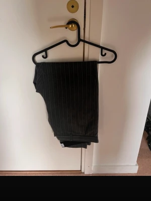 Randiga kostymbyxor från Just Junkies XL - Snygga mörkgrå kostymbyxor från Just Junkies i storlek XL. Byxorna har tunna vita ränder som ger en fräsch och modern look. Klassisk rak passform med hällor för bälte och diskreta fickor. Perfekt för dig som gillar stilrena och trendiga plagg.