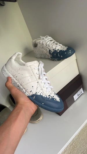Maison margiela - Unika sneakers i vitt och mörkblått med coolt färgstänksmönster över hela skon. Rund tå, platt sula och klassisk snörning. Skorna har en modern vibe och är tillverkade i skinn för en lyxig känsla. Perfekt för dig som vill sticka ut med din stil.