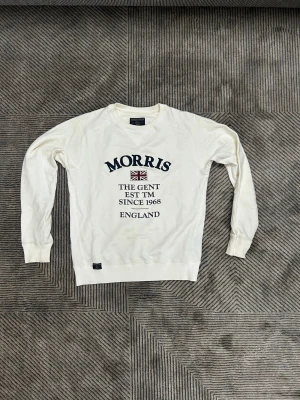 Vit sweatshirt från Morris  - Snygg vit sweatshirt från Morris Gents i storlek XS. Tröjan har tryck med text och brittisk flagga på bröstet, rund hals och långa ärmar. Tillverkad i mjuk bomull med ribbade muddar vid ärmslut och nederkant. Perfekt för en chill och stilren look.