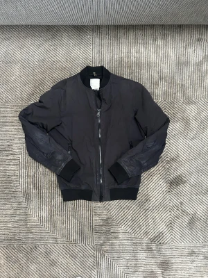 Svart bomberjacka J.Lindeberg S - Svart bomberjacka från J.Lindeberg med broderad bläckfisk och texten 'Arctic Black' på ryggen. Jackan har dragkedja framtill, ribbade muddar och meshfoder på insidan. Snygg och stilren med cool detalj på ryggen.