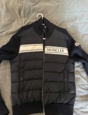 Svart Moncler pufferjacka med stickade ärmar - Säljer en svart pufferjacka från Moncler med stickade ärmar och rygg. Jackan har vit logga och rand över bröstet, hög krage och dragkedja framtill. Klassisk Moncler-patch på ena ärmen. Perfekt för dig som vill ha en snygg och sportig look. Priset kan diskuteras.