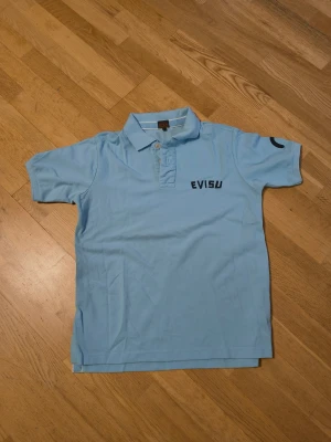 Ljusblå Evisu polotopp med tryck - Säljer en ljusblå polotopp från Evisu med korta ärmar och krage. Framsidan har Evisu-logga och baksidan har ett stort, färgglatt ansiktstryck med bling bling detaljer. Toppen är gjord i mjuk bomull och har knappar vid halsen. Perfekt för dig som gillar streetwear och unika prints.